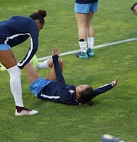 A group of athletes in sportswear are on a green soccer field. Two individuals are interacting closely; one helps the other who is lying on the grass. Nearby, other athletes are stretching.