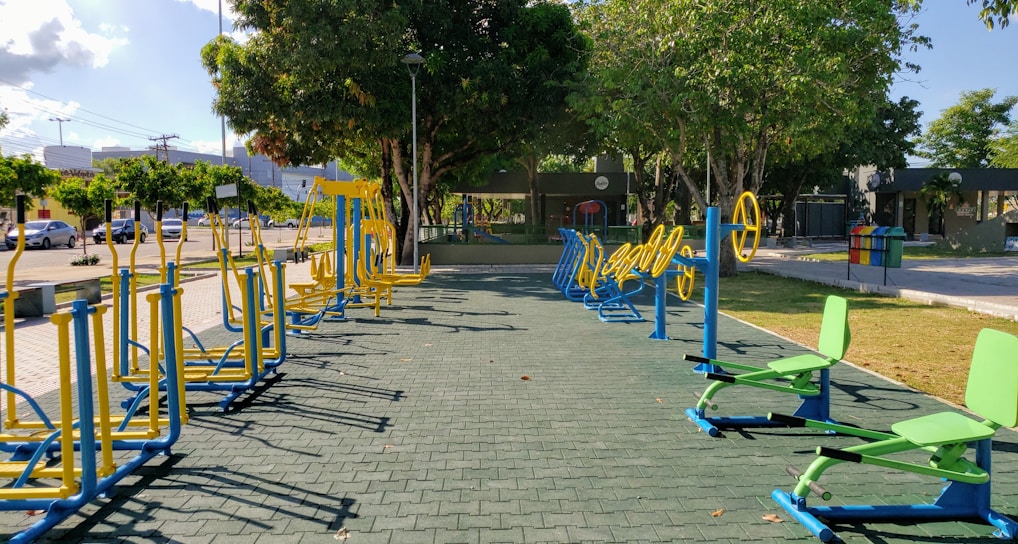 An outdoor fitness area featuring a variety of exercise equipment, including stationary bikes and other workout stations. The equipment is primarily blue and yellow, arranged neatly on a paved surface. Surrounding the area are trees providing shade, with a few parked cars and buildings visible in the background.