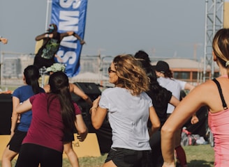 A group of people appears to be participating in an outdoor fitness class or dance session. They are dressed in athletic clothing, and a blue banner can be seen in the background, along with a stage. The setting suggests a lively and energetic atmosphere.