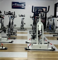 A room filled with stationary exercise bikes arranged neatly in rows. The space has a wooden and gray tiled floor, and a sign with the text 'i-Fitness' is displayed on the wall. The bikes are primarily white with black handlebars and seats.