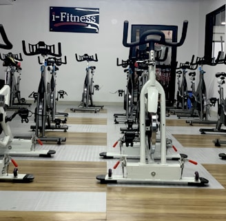 A room filled with stationary exercise bikes arranged neatly in rows. The space has a wooden and gray tiled floor, and a sign with the text 'i-Fitness' is displayed on the wall. The bikes are primarily white with black handlebars and seats.