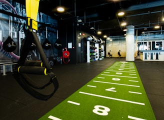 A modern gym space featuring a green track laid out on the floor with numbered lanes. TRX suspension trainers hang prominently in the foreground. The gym is equipped with various exercise equipment, including kettlebells and punching bags. In the background, there is a FIT sign near a reception desk under a low-ceilinged industrial design.