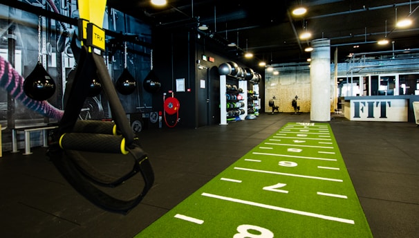 A modern gym space featuring a green track laid out on the floor with numbered lanes. TRX suspension trainers hang prominently in the foreground. The gym is equipped with various exercise equipment, including kettlebells and punching bags. In the background, there is a FIT sign near a reception desk under a low-ceilinged industrial design.