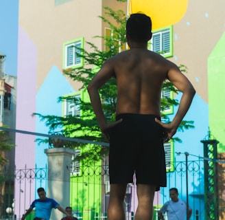 A group of people are engaged in a fitness activity outdoors. In the foreground, a man stands with his hands on his hips, facing away from the camera. In the background, other individuals are stretching or performing exercises. The setting is in front of a colorful, mural-covered building, with bright geometric patterns on the walls. A fence with decorative metalwork adds structure to the scene.