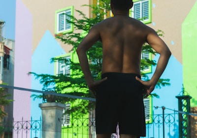 A group of people are engaged in a fitness activity outdoors. In the foreground, a man stands with his hands on his hips, facing away from the camera. In the background, other individuals are stretching or performing exercises. The setting is in front of a colorful, mural-covered building, with bright geometric patterns on the walls. A fence with decorative metalwork adds structure to the scene.