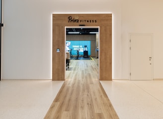 A modern fitness center entrance with wooden framing and the sign 'PRIME FITNESS' above the doorway. The interior features a spacious gym area with workout equipment visible and a motivational banner on the back wall. The flooring transitions from glossy tiles to a wood finish leading into the facility.