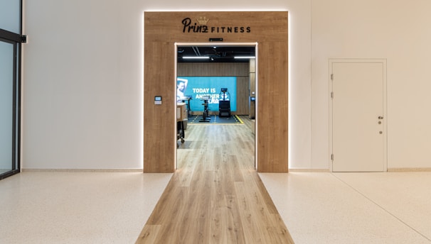 A modern fitness center entrance with wooden framing and the sign 'PRIME FITNESS' above the doorway. The interior features a spacious gym area with workout equipment visible and a motivational banner on the back wall. The flooring transitions from glossy tiles to a wood finish leading into the facility.