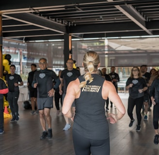 A group of individuals in athletic wear participate in a fitness class inside a spacious, well-lit room. The instructor, facing away, leads the group with energy and focus. The participants appear engaged and attentive, following along with the instructor's movements.