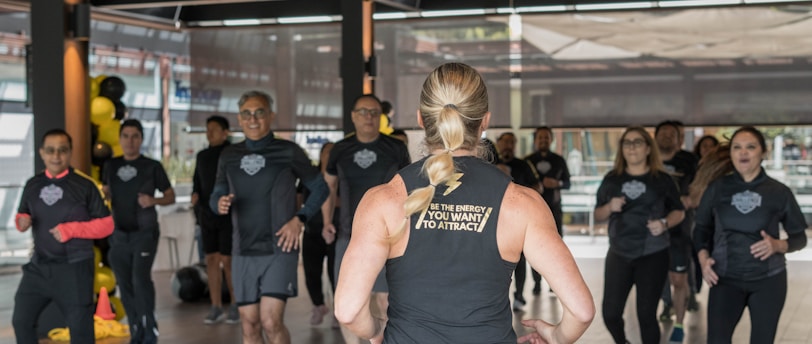 A group of individuals in athletic wear participate in a fitness class inside a spacious, well-lit room. The instructor, facing away, leads the group with energy and focus. The participants appear engaged and attentive, following along with the instructor's movements.