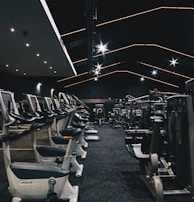 A spacious gym filled with various exercise equipment. Treadmills are aligned in rows on one side, while weight machines and other fitness devices are organized on the other side. The ceiling features modern, angular lines with bright overhead lights, adding a sleek and contemporary feel.