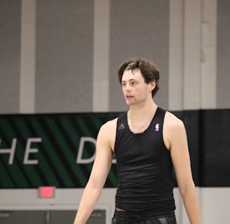A person wearing a sleeveless black sports top is standing indoors in a gymnasium. The background includes a gray wall with green and white lettering partially visible.