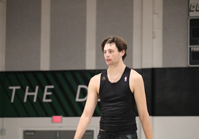 A person wearing a sleeveless black sports top is standing indoors in a gymnasium. The background includes a gray wall with green and white lettering partially visible.
