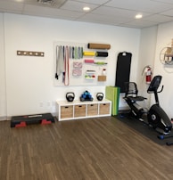 A fitness room featuring a variety of workout equipment neatly organized. The wall has a pegboard displaying exercise bands, foam rollers, and ropes. Below the board, there are kettlebells and dumbbells on shelves. An exercise bike is positioned to the right, with a step platform on the left.