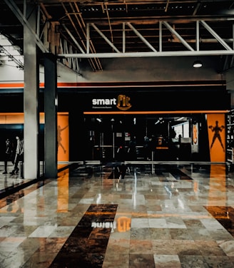 A fitness center entrance with the branding 'smart fit' illuminated above the doors. The interior has a modern design, featuring dark glass and bright orange accents. A reflective marble floor extends in front, capturing reflections from the surroundings. There are two individuals standing and walking towards the left.