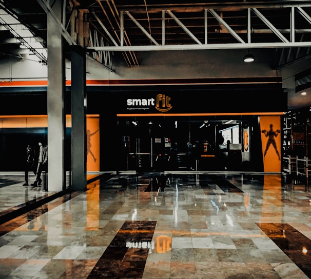 A fitness center entrance with the branding 'smart fit' illuminated above the doors. The interior has a modern design, featuring dark glass and bright orange accents. A reflective marble floor extends in front, capturing reflections from the surroundings. There are two individuals standing and walking towards the left.