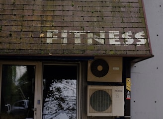 A building entrance with a door and windows is visible. Above the door, there is a sloped roof with the word 'FITNESS' written on it in white letters. An air conditioning unit is mounted next to the door. Leaves are scattered on the ground, and tree branches with leaves hang over the roof.