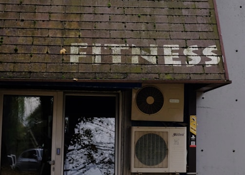 A building entrance with a door and windows is visible. Above the door, there is a sloped roof with the word 'FITNESS' written on it in white letters. An air conditioning unit is mounted next to the door. Leaves are scattered on the ground, and tree branches with leaves hang over the roof.